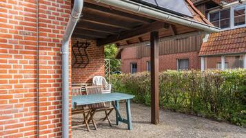 Een overdekt terras met een tafel en stoelen, een bakstenen muur en een haag.