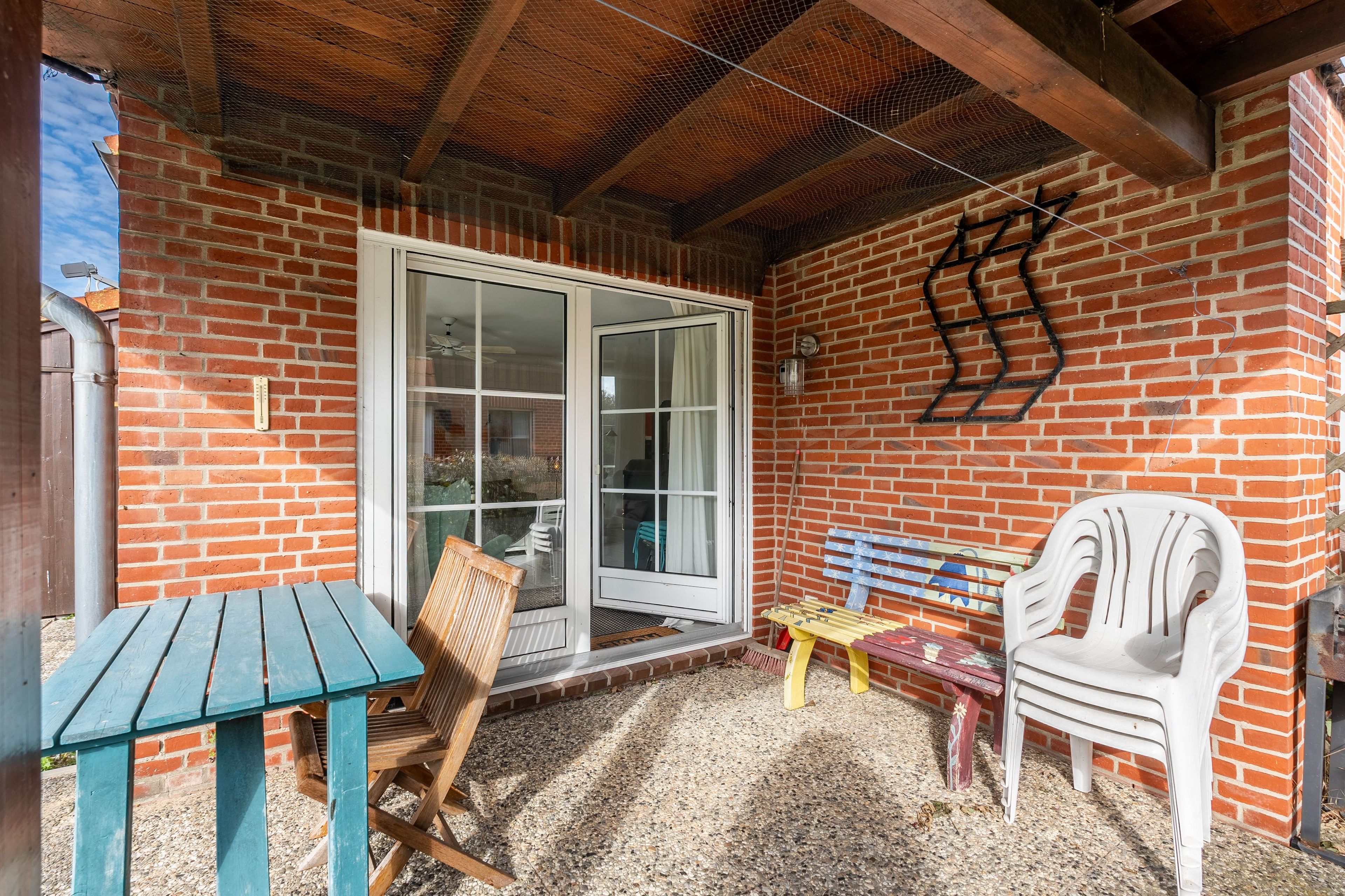 Un patio couvert avec un mur en briques, une porte-fenêtre coulissante, une table en bois, un banc et une chaise en plastique blanche.
