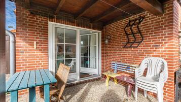 Un patio couvert avec un mur en briques, une porte-fenêtre coulissante, une table en bois, un banc et une chaise en plastique blanche.