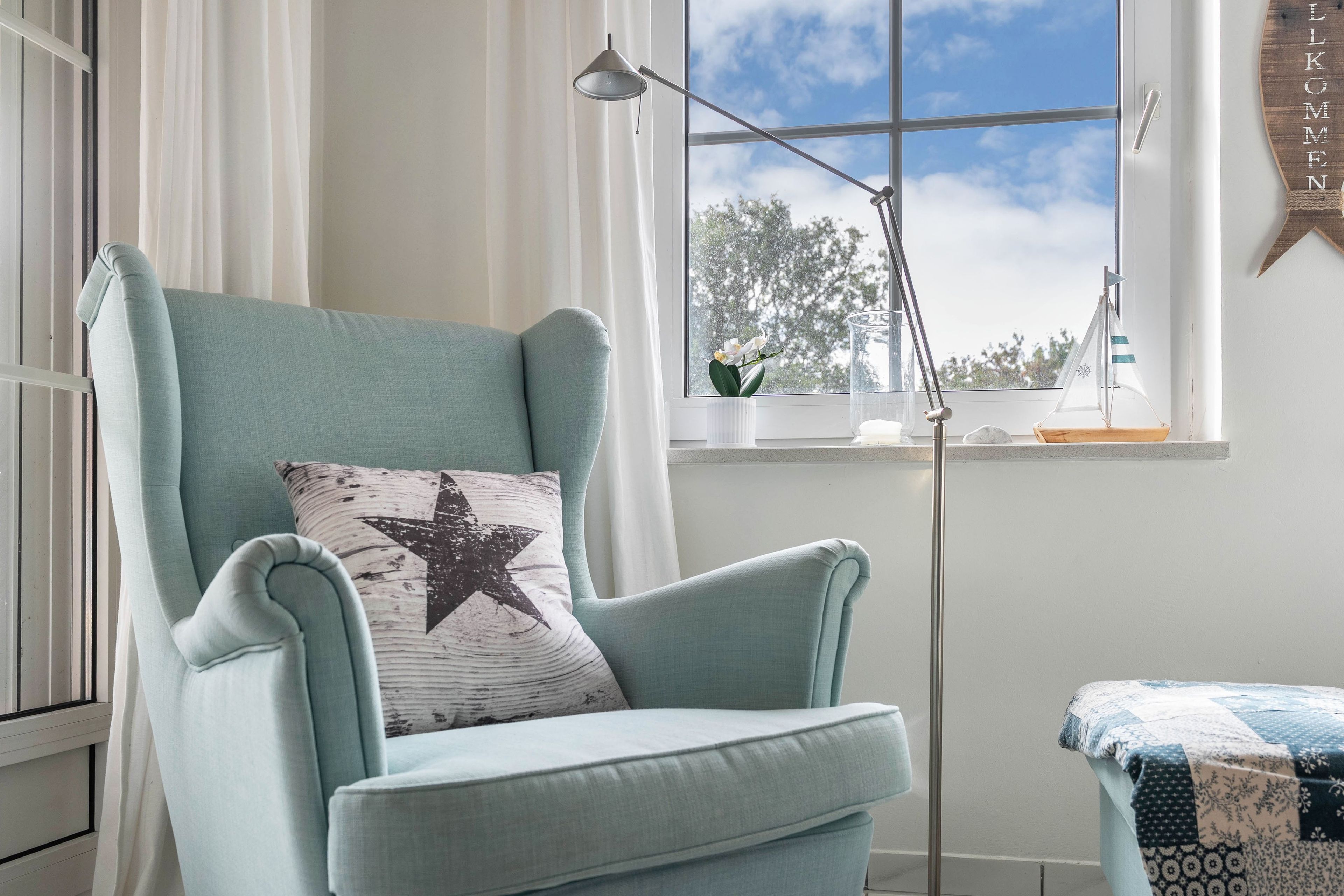 Ein hellblauer Sessel mit Kopfkissen in Form eines Sterns, ein kleiner Tisch mit einer Decke, eine Stehlampe und ein Fenster mit Blick auf Bäume und Himmel.