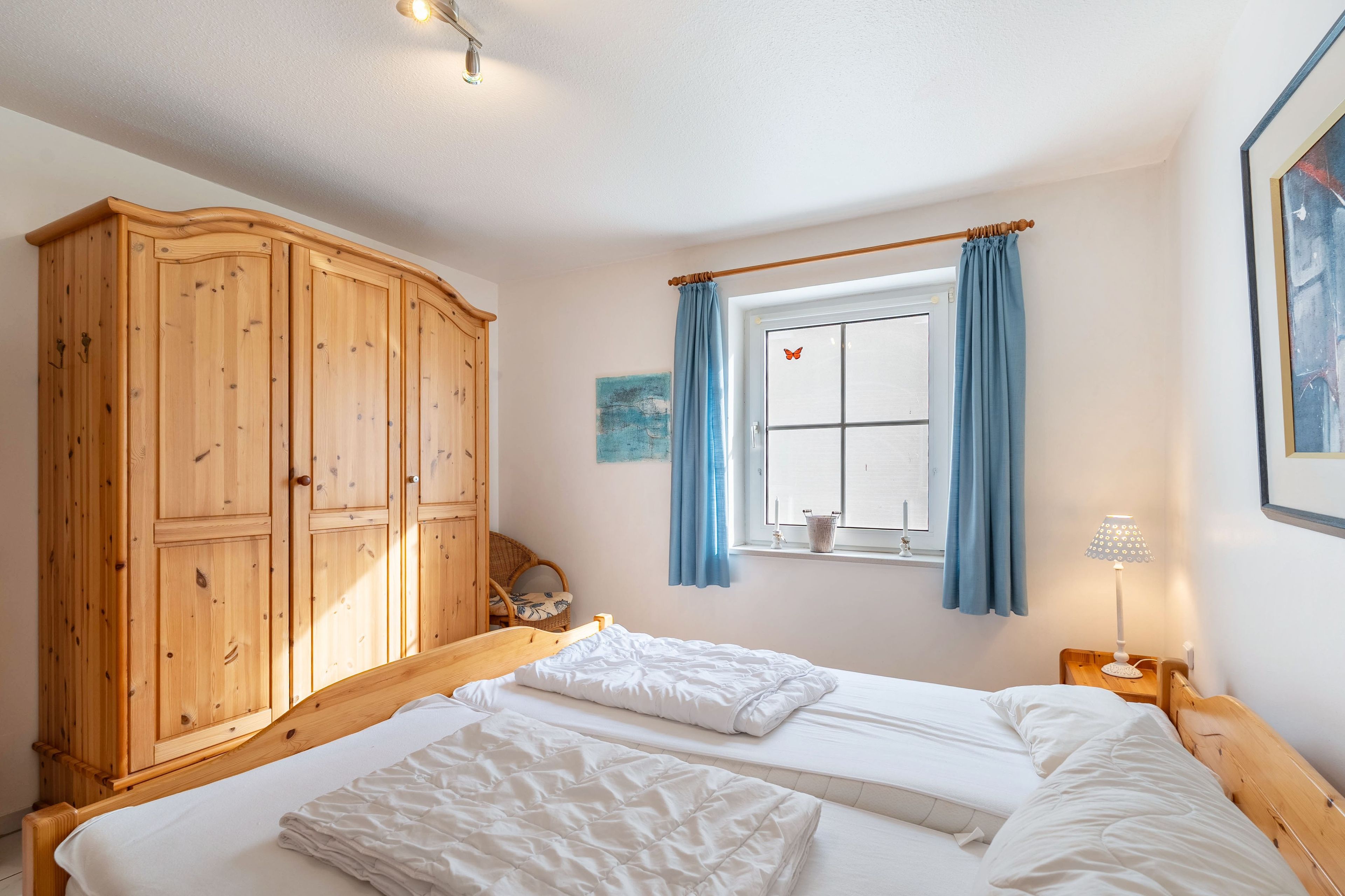 Une chambre avec une armoire en bois, un lit avec une literie blanche, une fenêtre avec des rideaux bleus et une table de chevet avec une lampe.