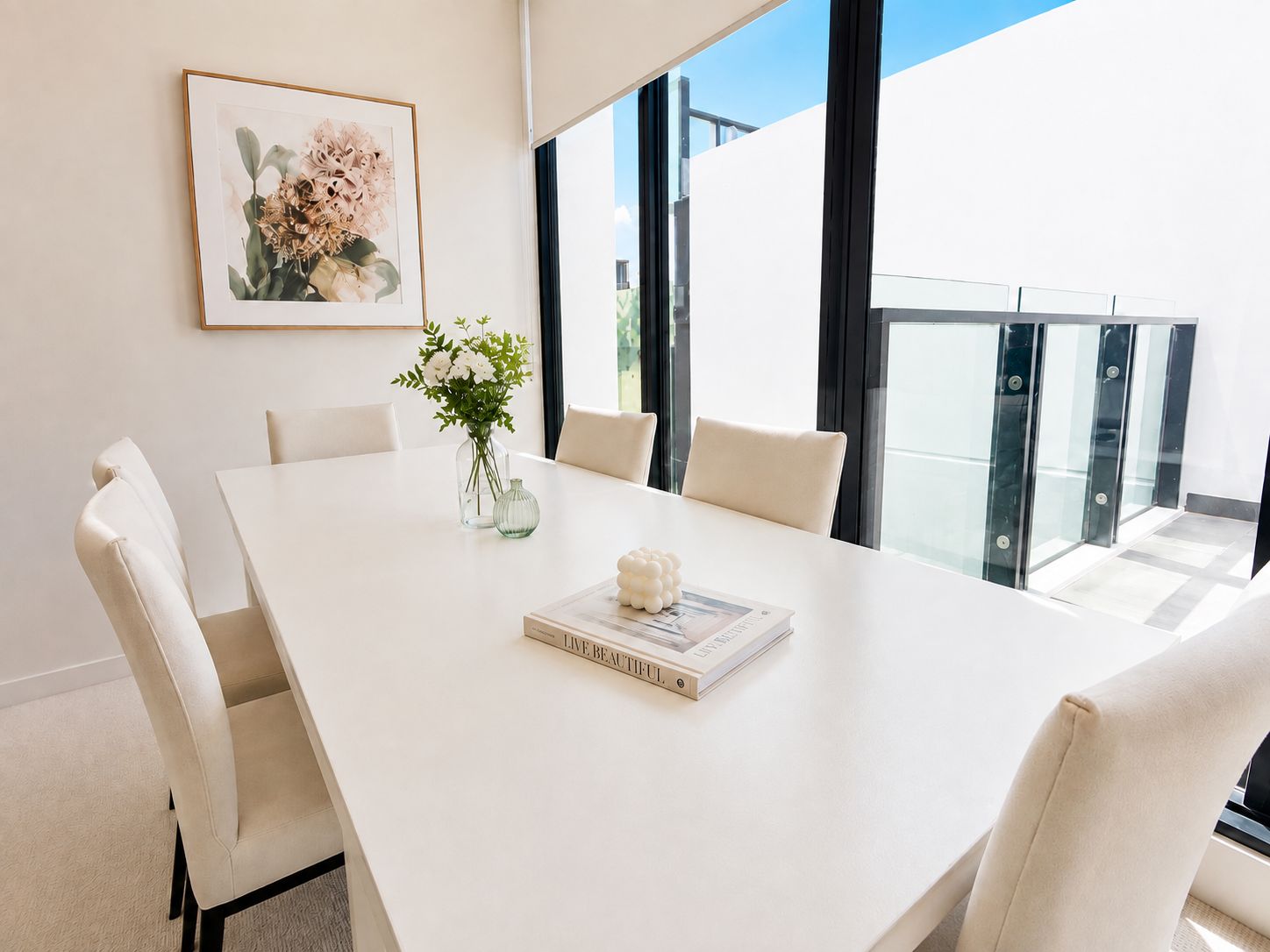 Une salle à manger moderne avec une table blanche, six chaises, un vase de fleurs et un tableau encadré au mur.
