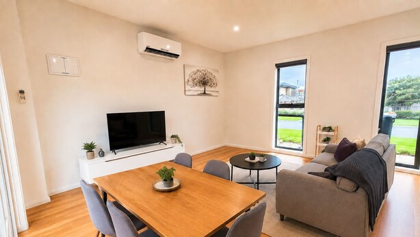 A modern living room with a wooden dining table, grey sofas, a flat-screen TV, and a large window offering a view of the outdoors. - The Glenroy Getaway – 2BR Parking & Wi-Fi (Glenroy)