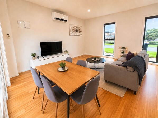 Una sala de estar moderna con una mesa de comedor de madera, sofás grises, un televisor de pantalla plana y un amplio ventanal con vistas al exterior.