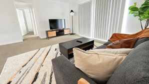 Uma sala de estar moderna com sofá, mesa de centro, TV em móvel e uma grande janela com persianas.