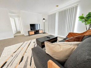 A modern living room with a sofa, a coffee table, a TV on a stand, and a large window with blinds. - Elegant Lakeside Escape | Spacious 4BR Home Parkin (Point cook)