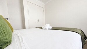 Uma cama de solteiro com colchão branco, manta verde, travesseiro branco e uma toalha branca enrolada na cama.