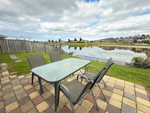A patio with a table and chairs overlooking a pond, surrounded by a wooden fence and houses in the background. - Elegant Lakeside Escape | Spacious 4BR Home Parkin (Point cook)