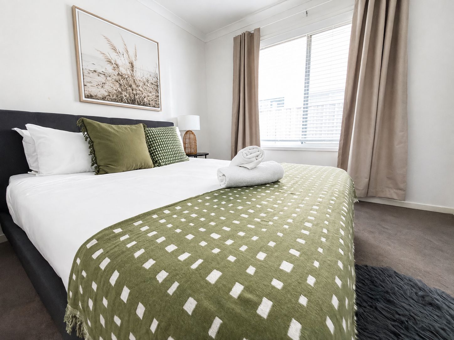 A hotel room with a bed, a window with curtains, a framed picture on the wall, and a nightstand with a lamp.