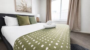 A hotel room with a bed, a window with curtains, a framed picture on the wall, and a nightstand with a lamp.