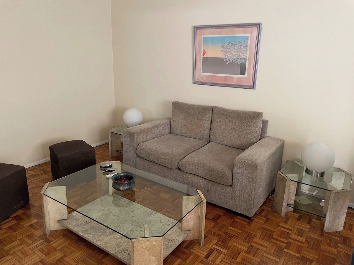 A living room with a glass coffee table, a grey sofa, a black armchair, a small side table with a lamp, and a framed picture on the wall.