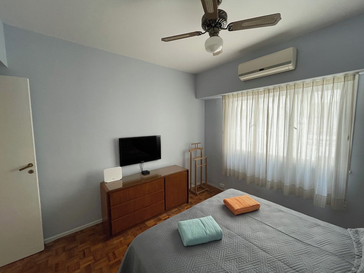A bedroom with a bed, a television on a wooden dresser, an air conditioning unit, and a window with sheer curtains.