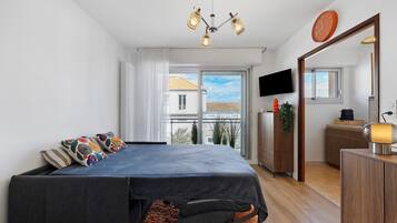 Une chambre avec un lit, une télévision, un miroir et un balcon donnant sur des immeubles.