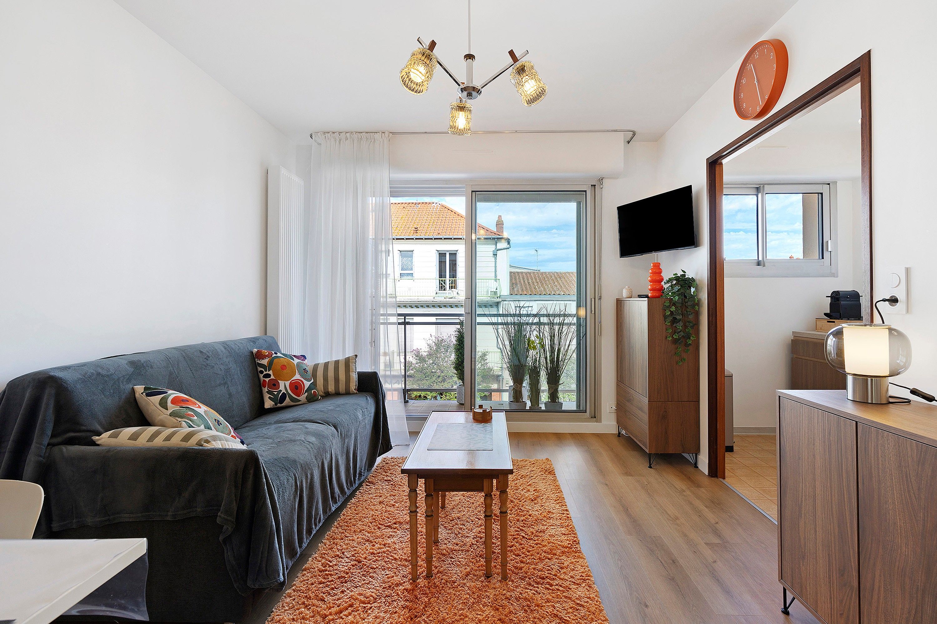 En moderne stue med sofa, bord, fjernsyn og en balkon med udsigt til bygninger.