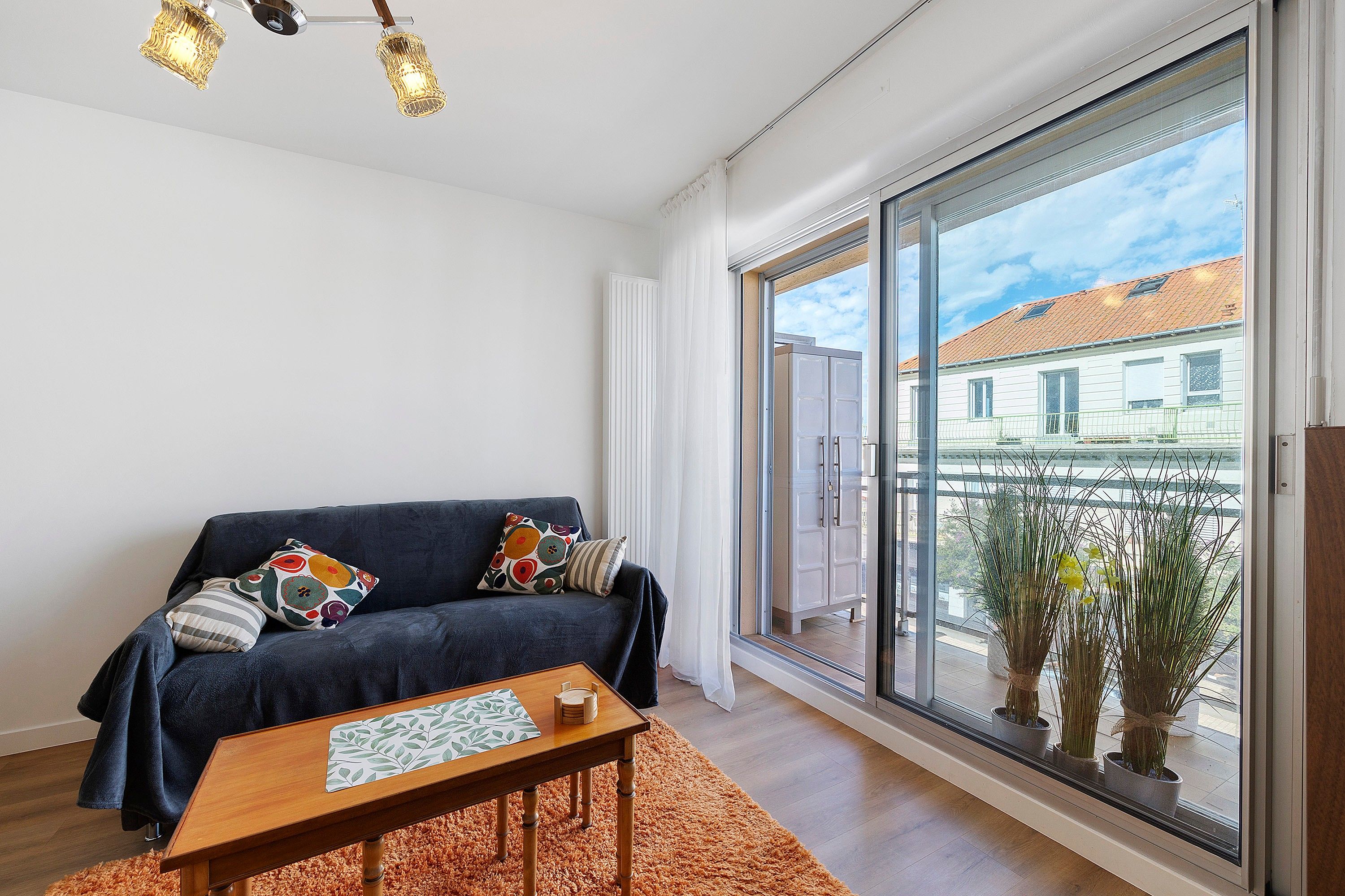 A modern living room with a dark blue sofa, patterned cushions, a wooden coffee table, and a large sliding glass door leading to a balcony with potted plants.