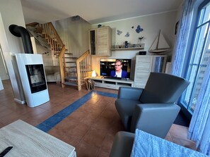 A living room with a TV, a sofa, a fireplace, a staircase, and a window with curtains. - "Sommerwind" townhouse with 95 square meters of living space and a large, quiet lot (fenced), close to downtown (Zingst)