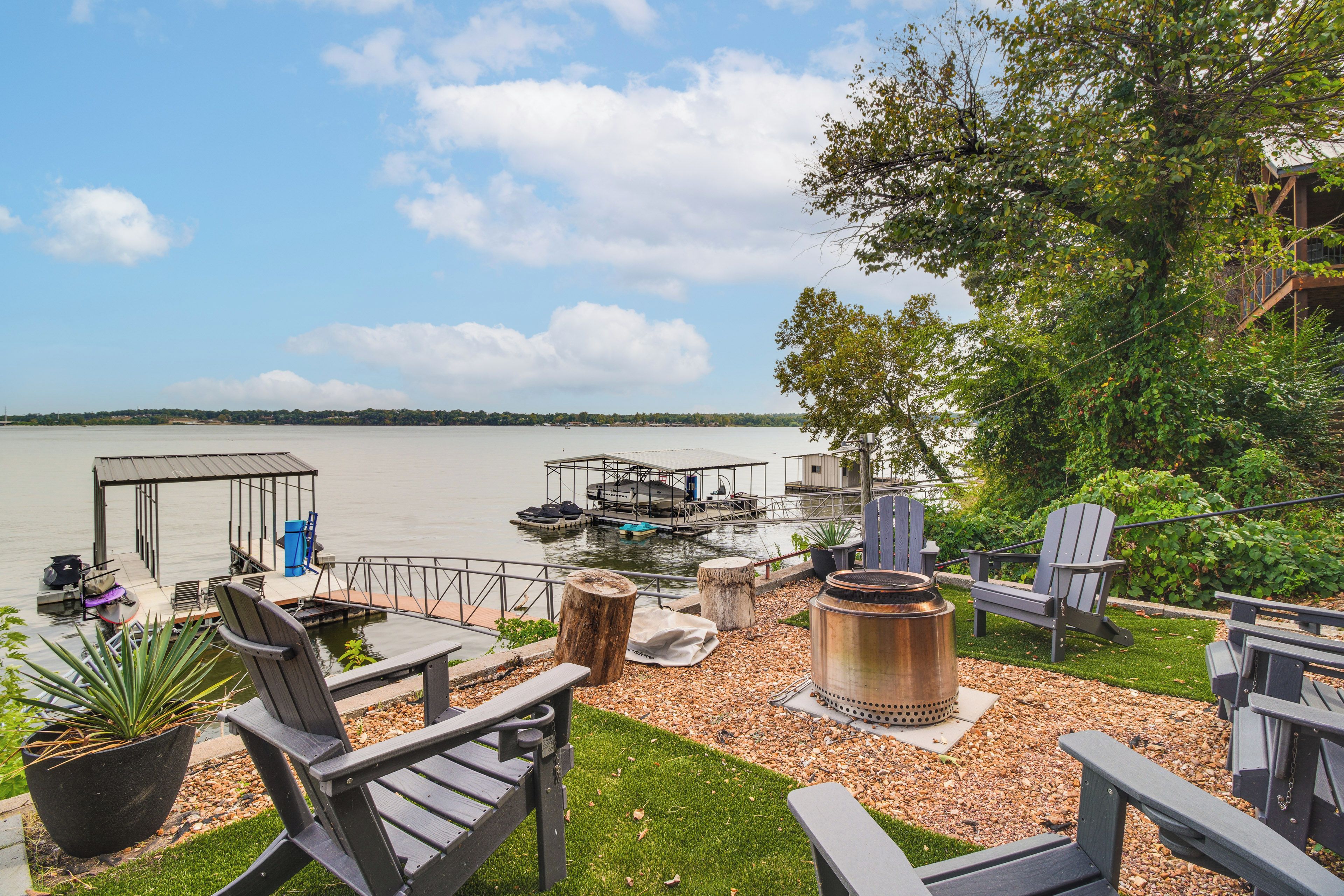 A lakeside deck with Adirondack chairs, a fire pit, and a dock with boats.