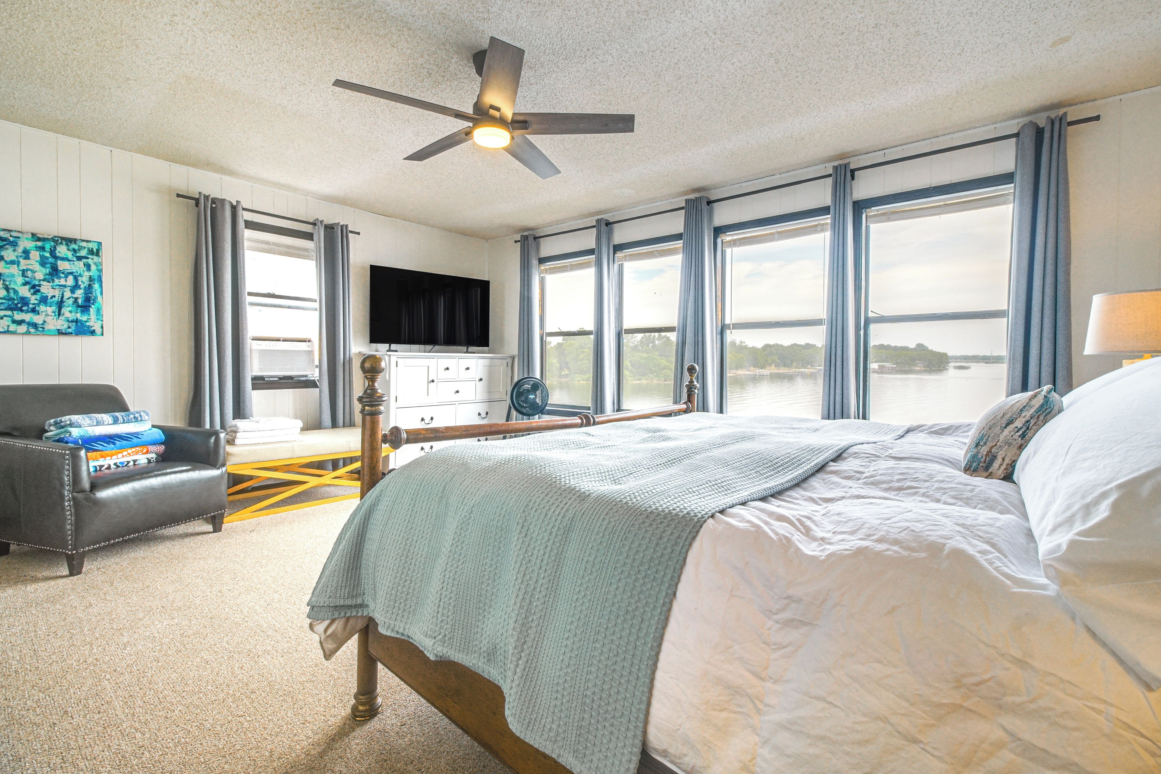 A bedroom with a bed, a desk, a chair, a television, and a view of a body of water through large windows.
