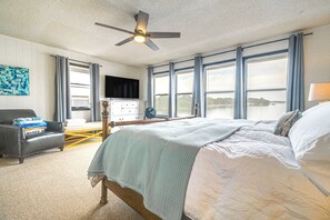Une chambre à coucher comprenant un lit, un bureau, une chaise, un téléviseur et offrant une vue sur une étendue d’eau grâce à de grandes fenêtres.