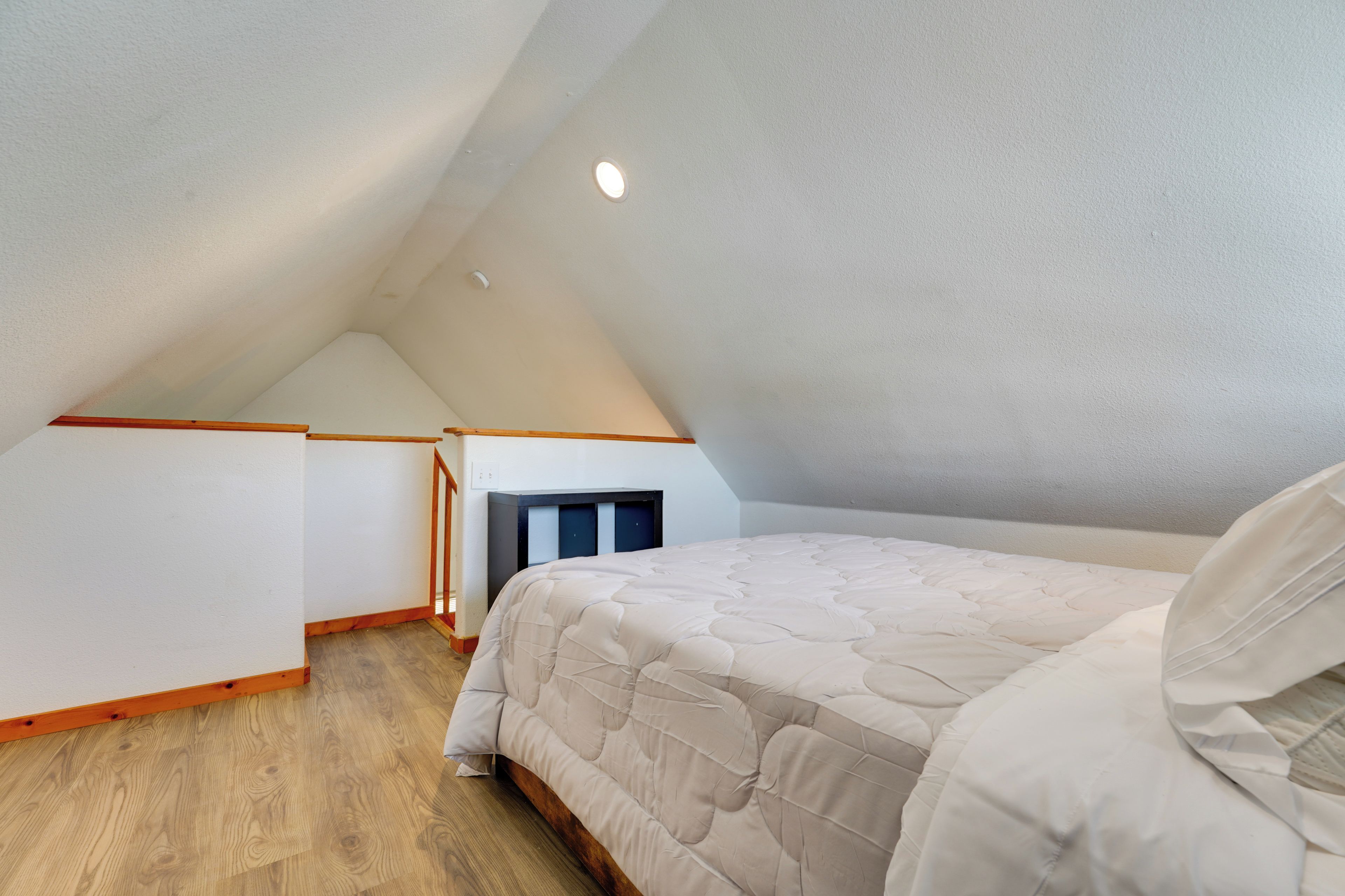 Quarto com teto inclinado, cama com edredom branco, piso de madeira e pequena lareira.