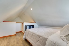 A bedroom with a sloped ceiling, a bed with a white duvet, wooden flooring, and a small fireplace.
