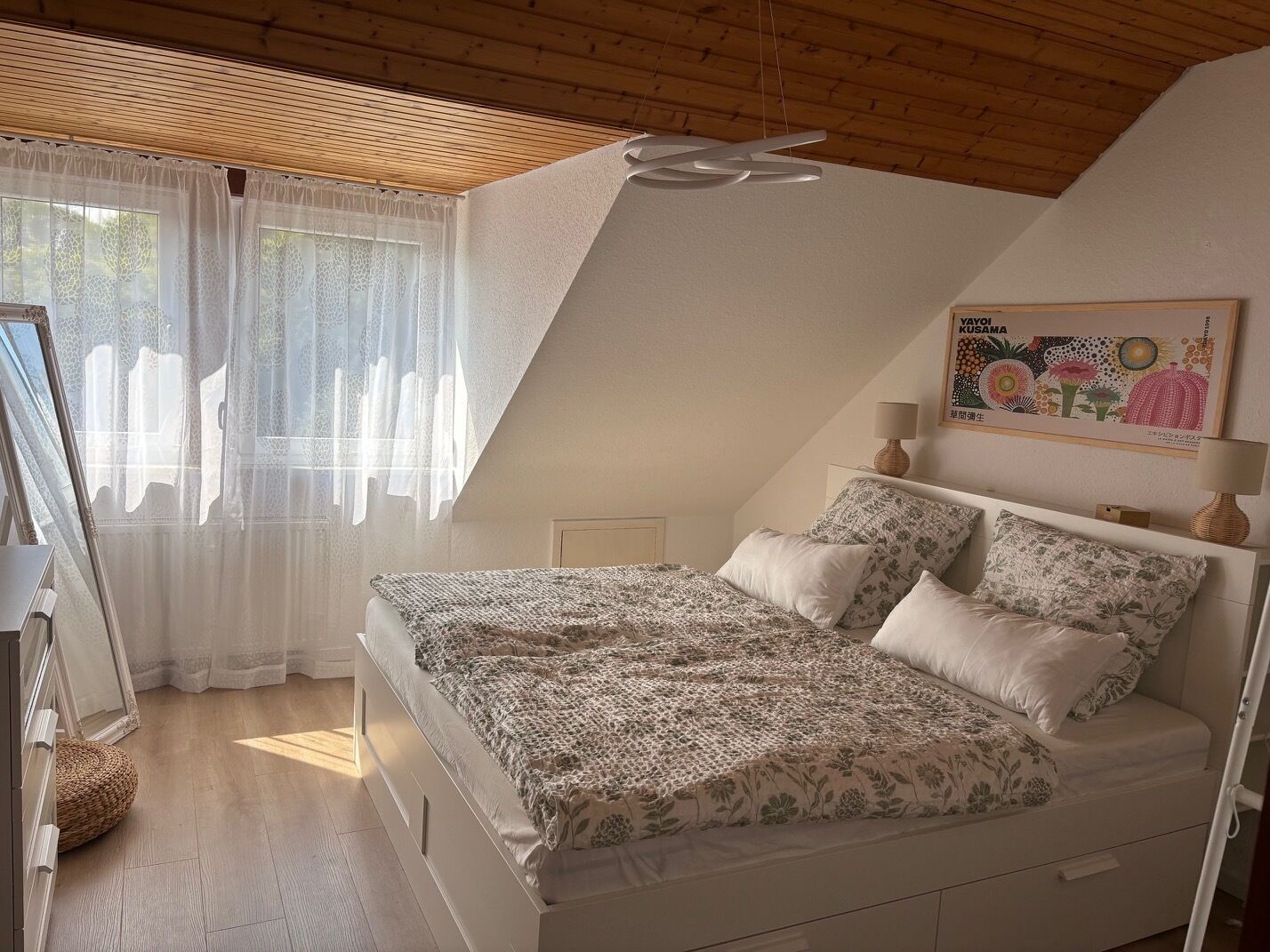 Ein Schlafzimmer mit schrägen Wänden, einem Bett mit gemusterten Betttextilien, einem Fenster mit transparenten Vorhängen und einer Wandleuchte.
