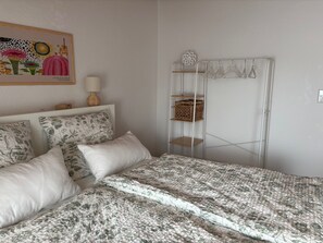 A bedroom with a bed, pillows, a bedside table, a lamp, a wall art piece, a wardrobe, and a hanging clothes rack.
