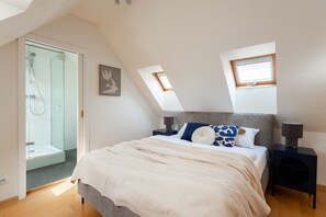 Quarto com cama, mesas de cabeceira, um espaço para objetos pessoais, janela e área de chuveiro.