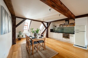 Un comedor con mesa y sillas de madera, una cocina con armarios y electrodomésticos blancos, y un techo inclinado con vigas a la vista.
