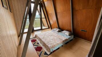 Quarto com teto inclinado, cama com roupa de cama estampada e vista para o verde lá fora.