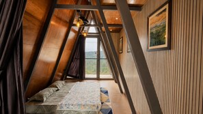 A room with a sloped ceiling, a bed with patterned bedding, a window with a view of greenery, and a framed picture on the wall. - Aara Villa (Mahabaleshwar)