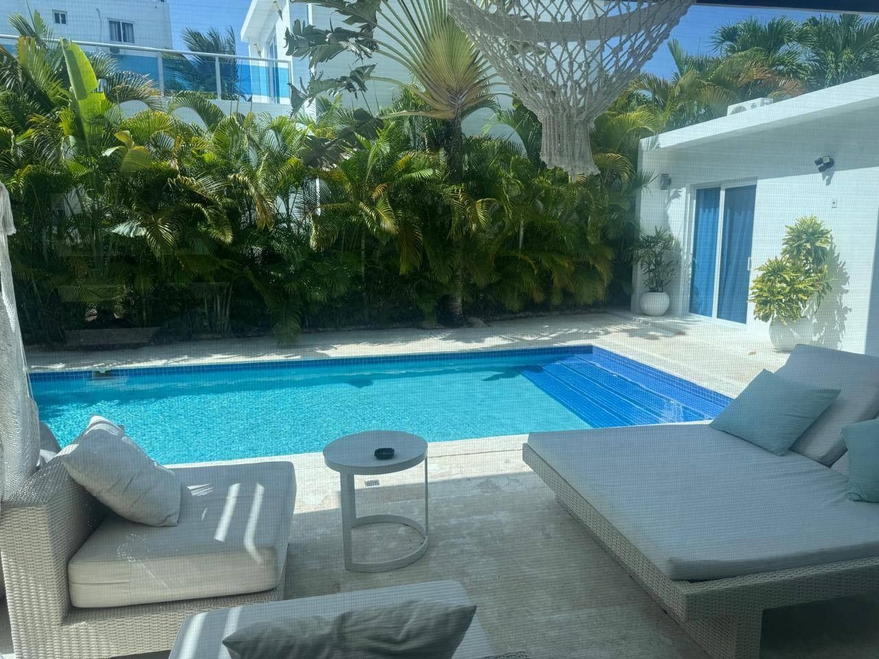 Ett område vid poolen med en solstol, ett litet runt bord och utsikt över en swimmingpool som omges av frodig grönska.