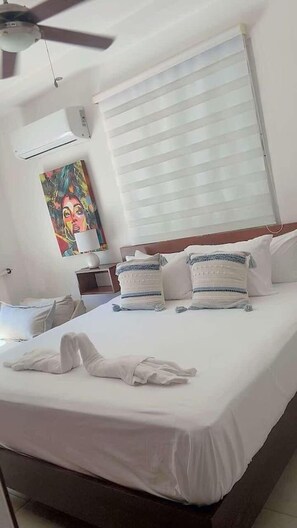 Ein Hotelzimmer mit einem Bett, einem Deckenventilator, einem Fenster mit Jalousien, einem Bild an der Wand und einem Nachttisch mit Lampe.