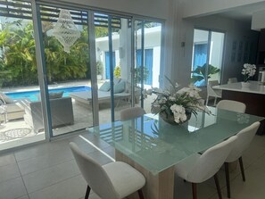 A dining area with a glass table and white chairs, a flower centerpiece, and a view of a pool and greenery outside. - Sosua Ocean Village - Las Palmas Villa #4 (Sosúa)