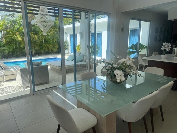 A dining area with a glass table and white chairs, a flower centerpiece, and a view of a pool and greenery outside. - Sosua Ocean Village - Las Palmas Villa #4 (Sosúa)