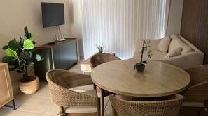 Een woonkamer met een ronde houten tafel, rieten stoelen, een bank, een televisie op een dressoir en een potplant.
