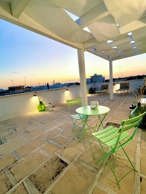 Eine Terrassesituation mit Tisch und Stühlen, einem grünen Blumentopf und Blick auf die Skyline der Stadt im Abenddämmerung.