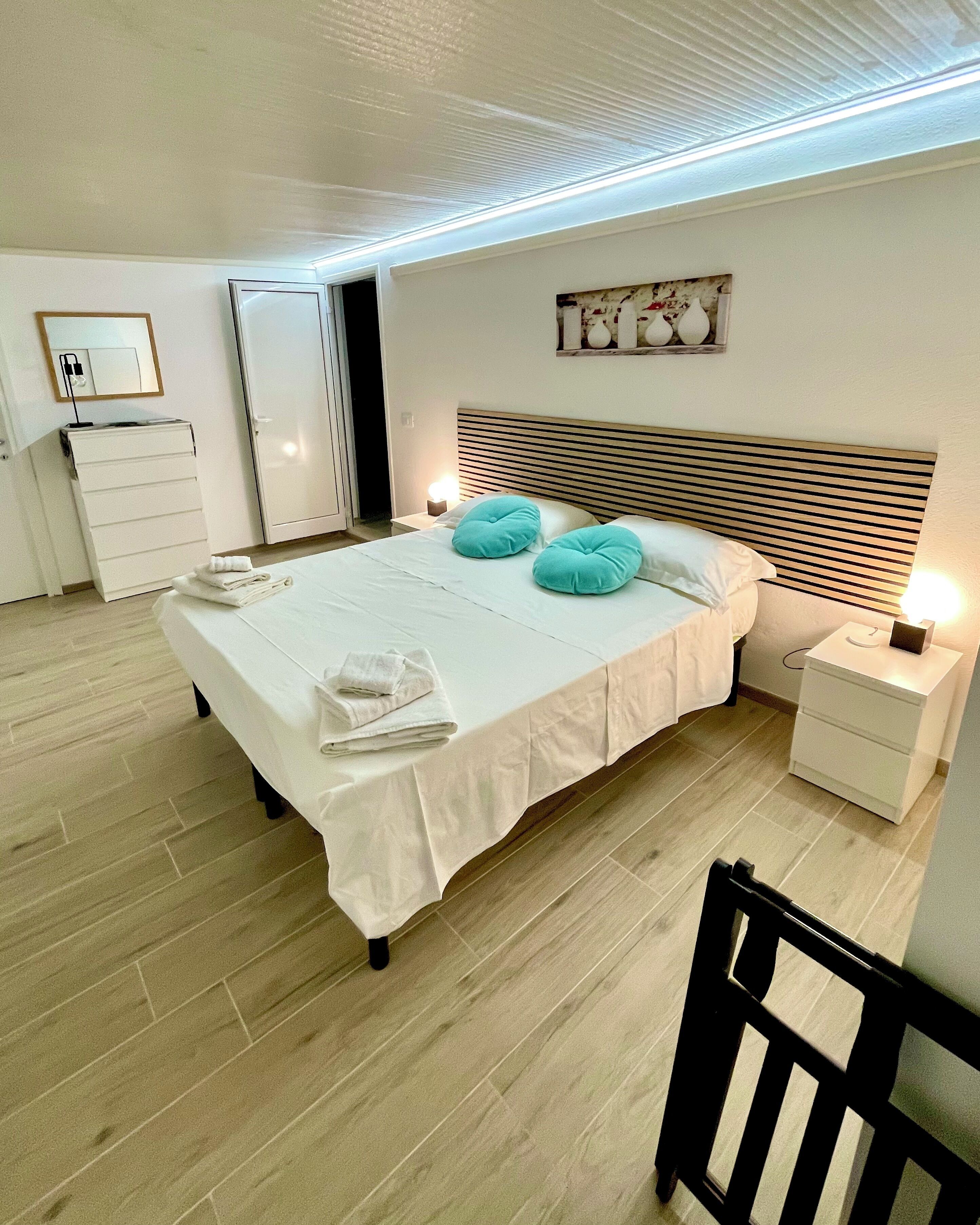 A neatly made bed with pillows and a folded towel, a bedside table with a lamp, and a chair in a room with wooden flooring and a slatted ceiling.