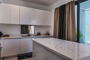 A modern kitchen with white cabinetry, a marble countertop, and a large window with curtains. - Poolside 2 Bedroom Apartment (Las Terrenas)