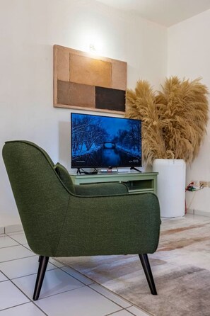 A modern living room with a green armchair, a flat-screen TV on a stand, a decorative wall piece, and a large potted plant. - One Bedroom Central Apartment (Sosúa)