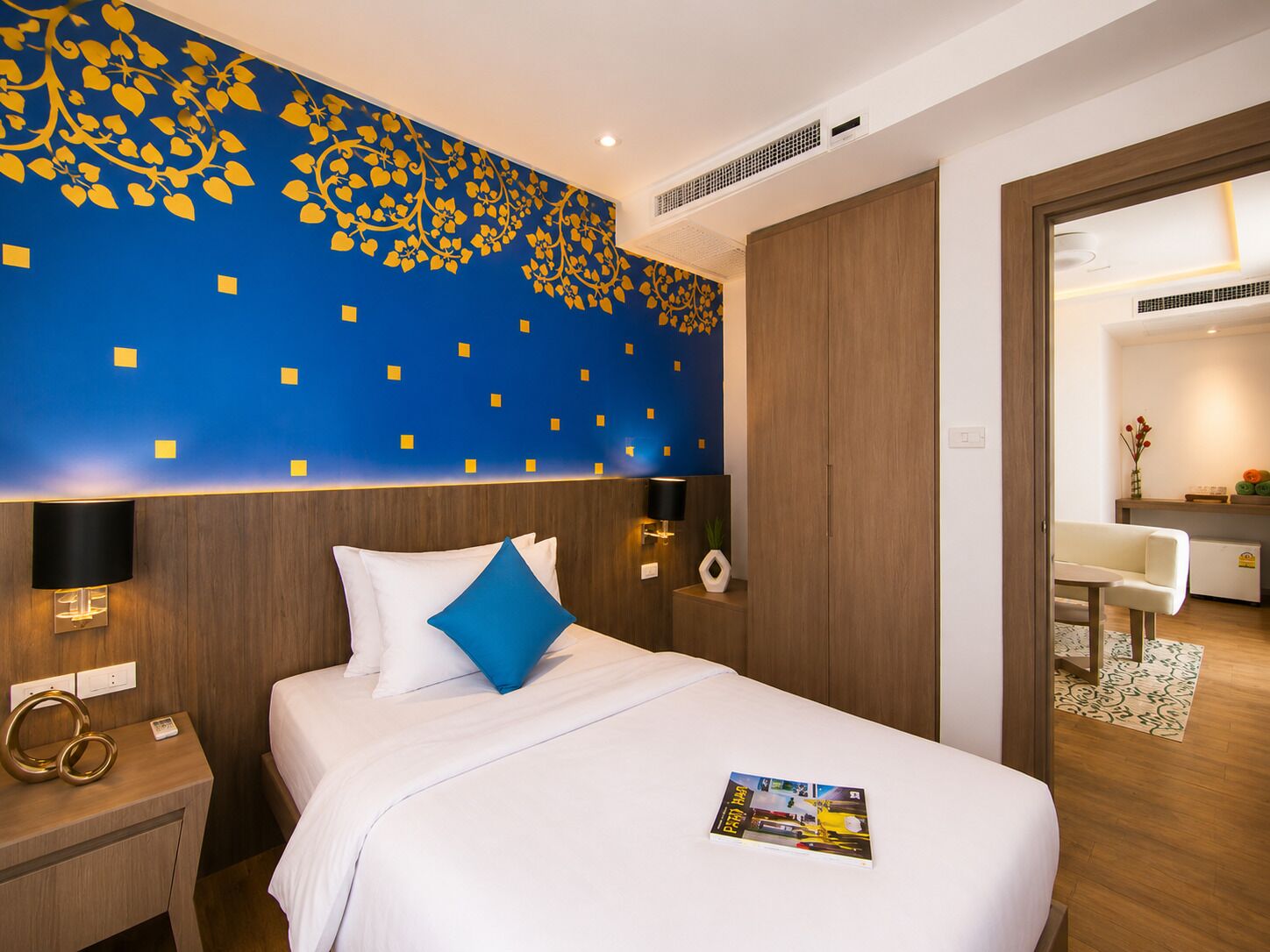 Ein Hotelzimmer mit Bett, Nachttisch, Wand mit Zierverzierungen und Blick in das angrenzende Zimmer.