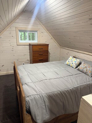 A bedroom with a sloped ceiling, a wooden bed frame, a bedside table, and a window. - Off the beaten path… (Algonquin Highlands)