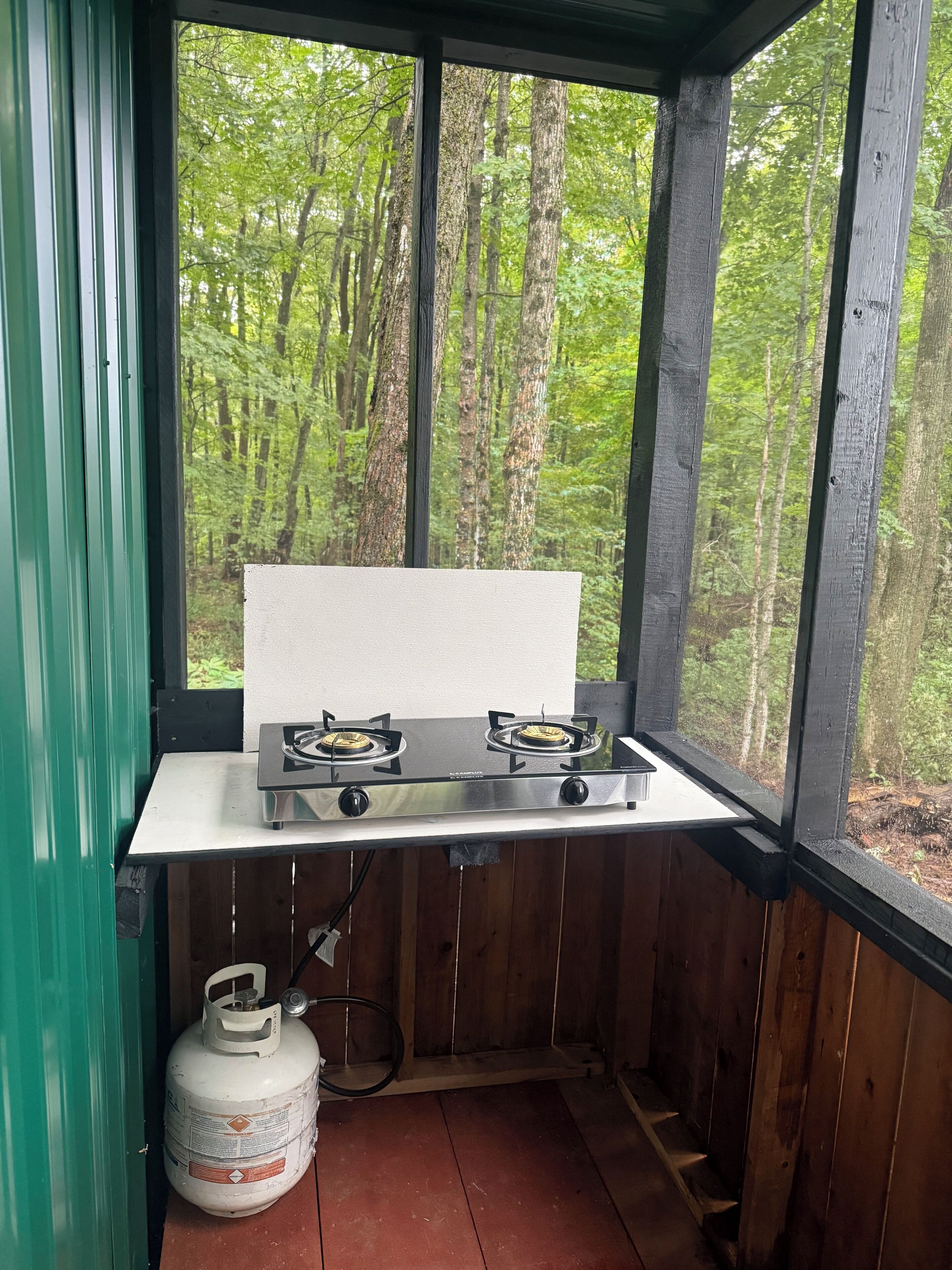 Une petite cuisine équipée d’une cuisinière à gaz et d’une bouteille de propane, donnant sur une forêt.