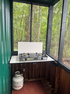 Une petite cuisine équipée d’une cuisinière à gaz et d’une bouteille de propane, donnant sur une forêt.