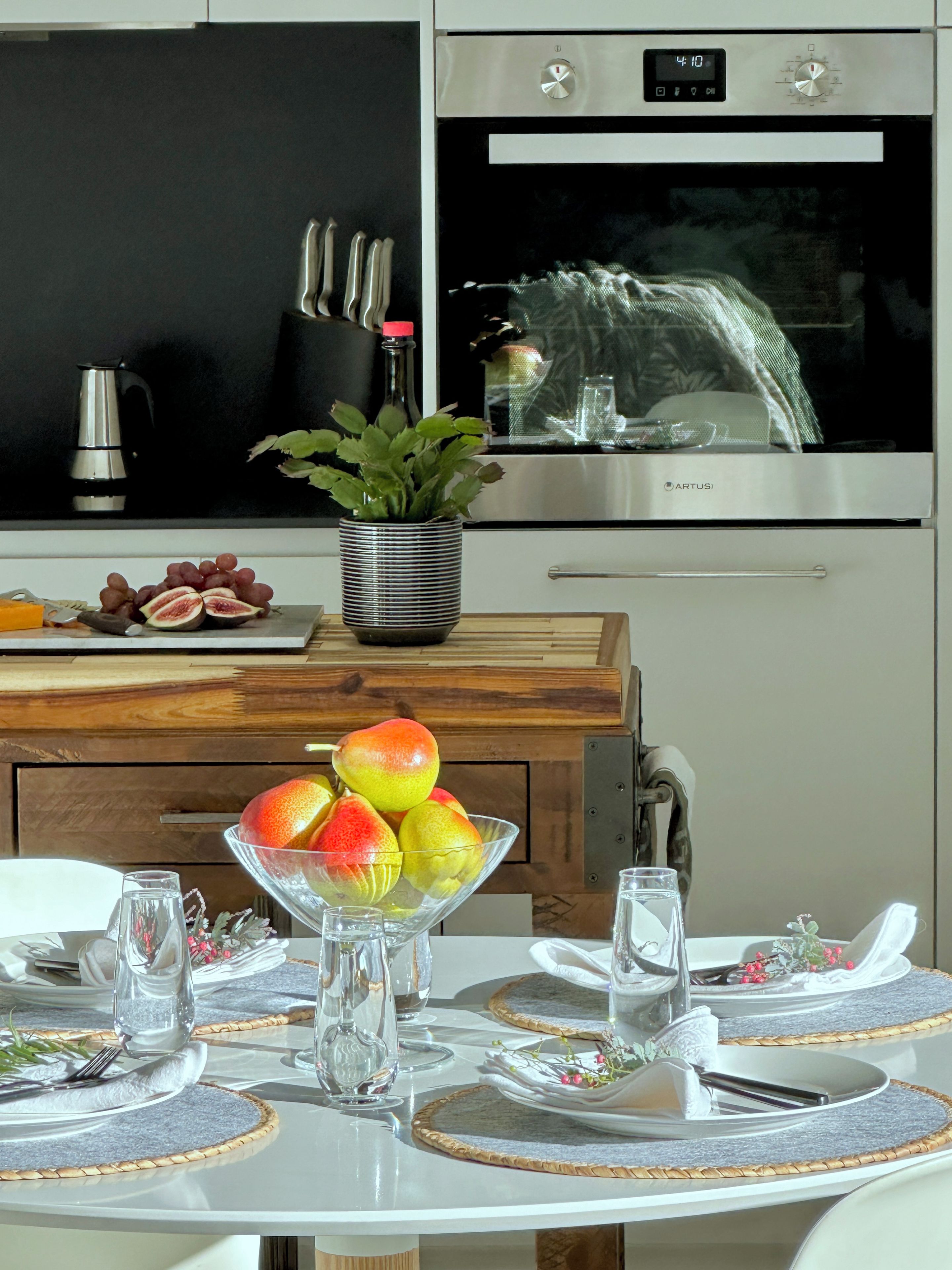 A modern kitchen with stainless steel appliances, a fruit bowl on the counter, and a dining area set with placemats and cutlery.