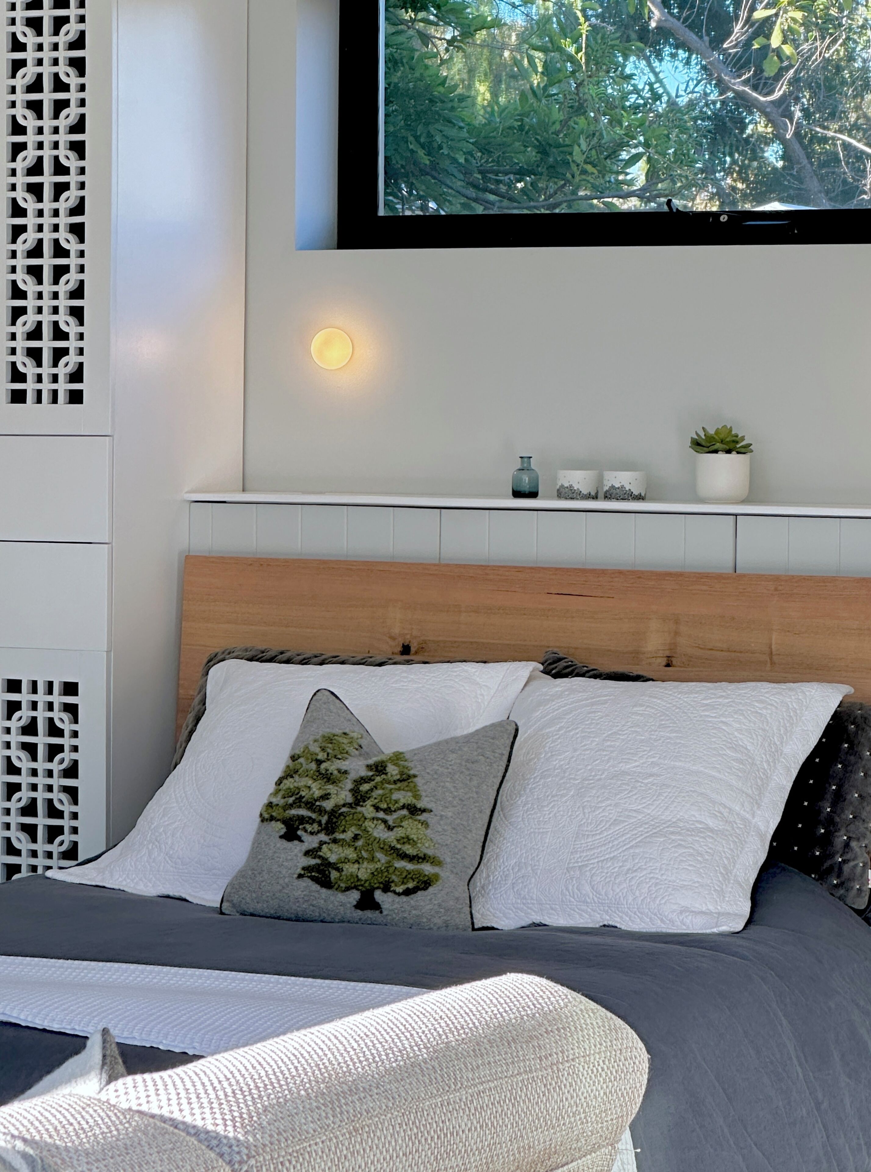 A bedroom with a bed, pillows, a bedside table, a wall-mounted TV, and a window with a view of trees.