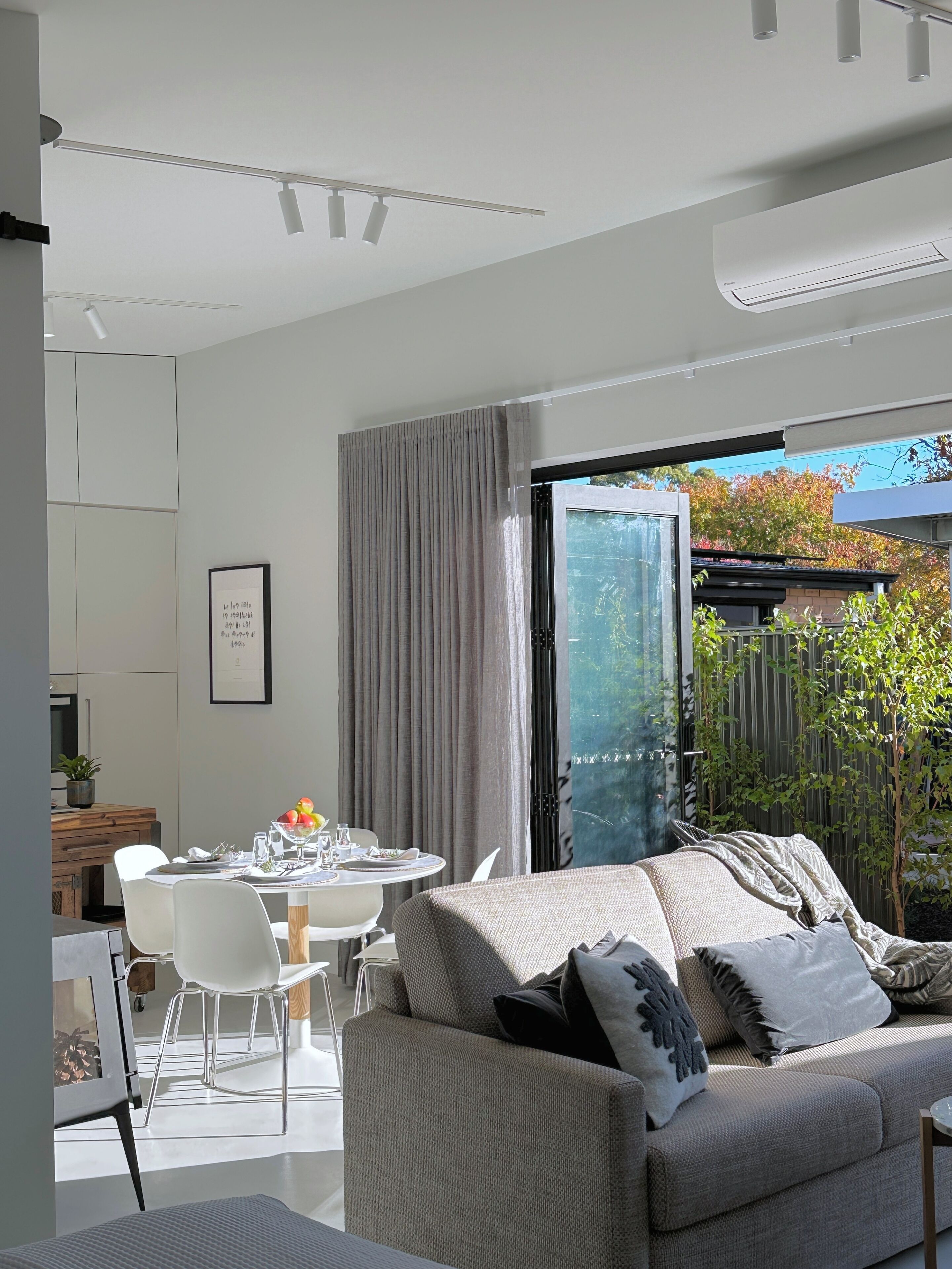 A modern living room with a sofa, dining table, and chairs. There is a sliding glass door leading to a garden.