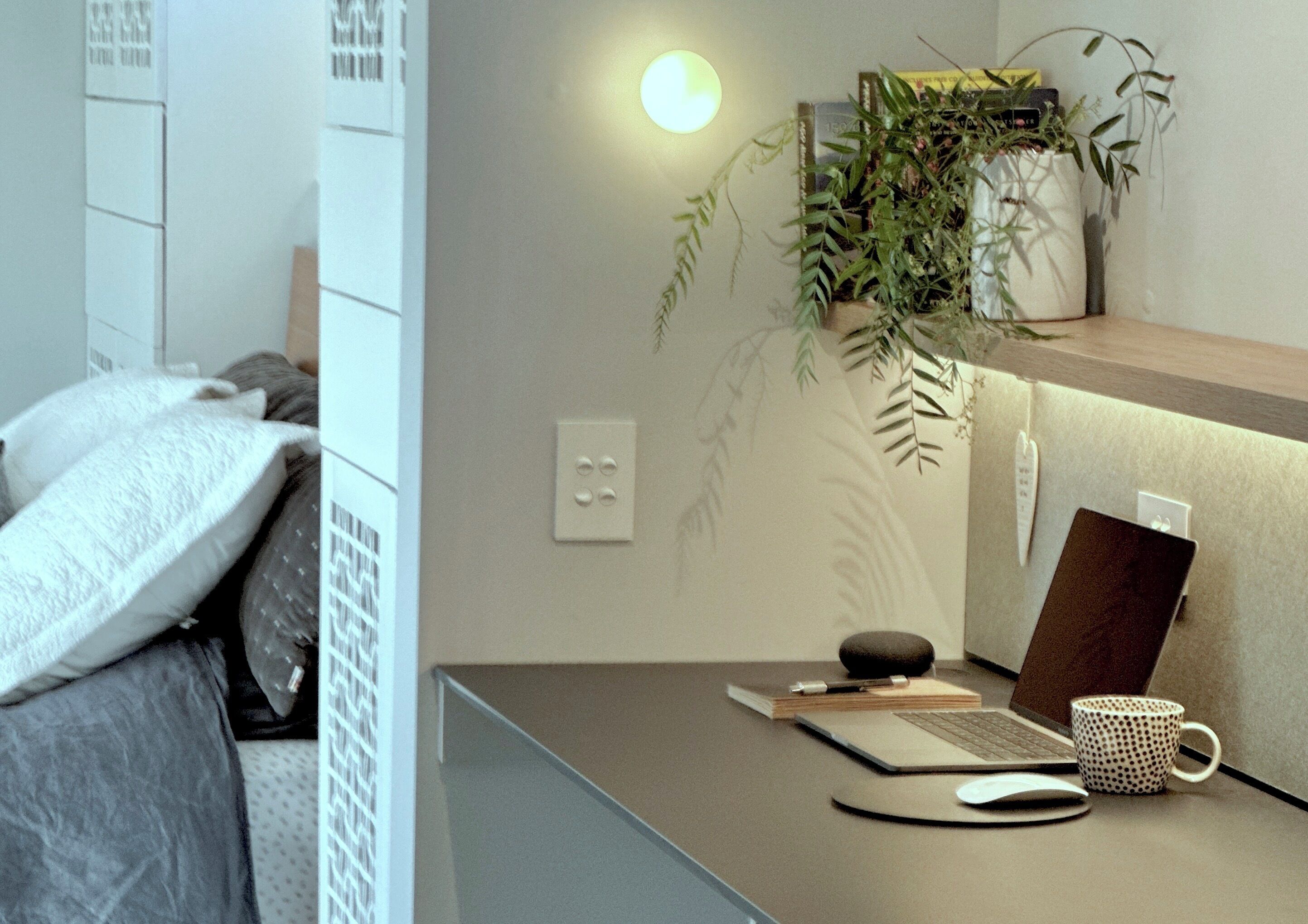 A bedroom with a bed, a bedside table with a lamp, and a wall-mounted shelf with a plant.