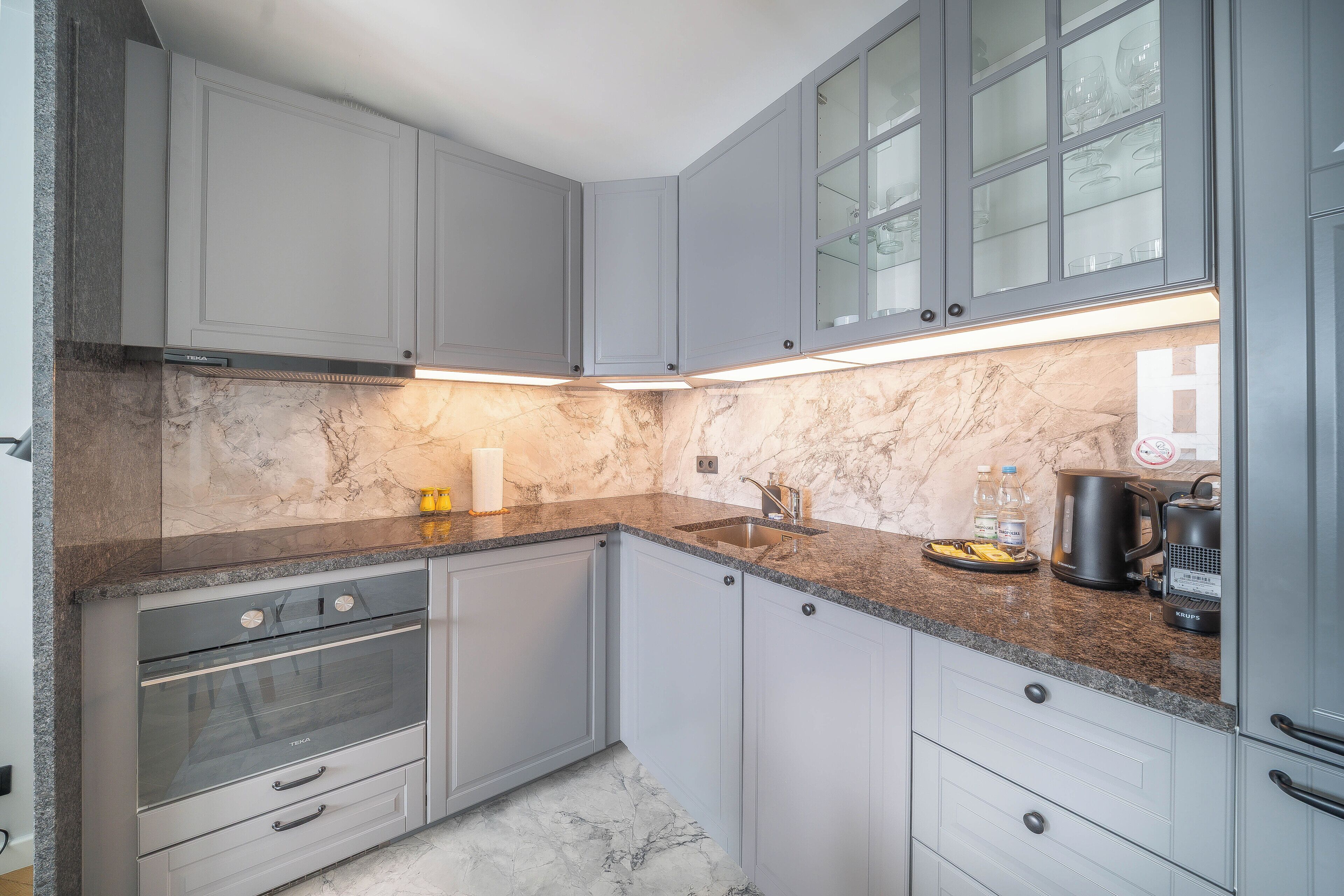 A modern kitchen with grey cabinets, a marble countertop, a built-in oven, and a coffee maker.