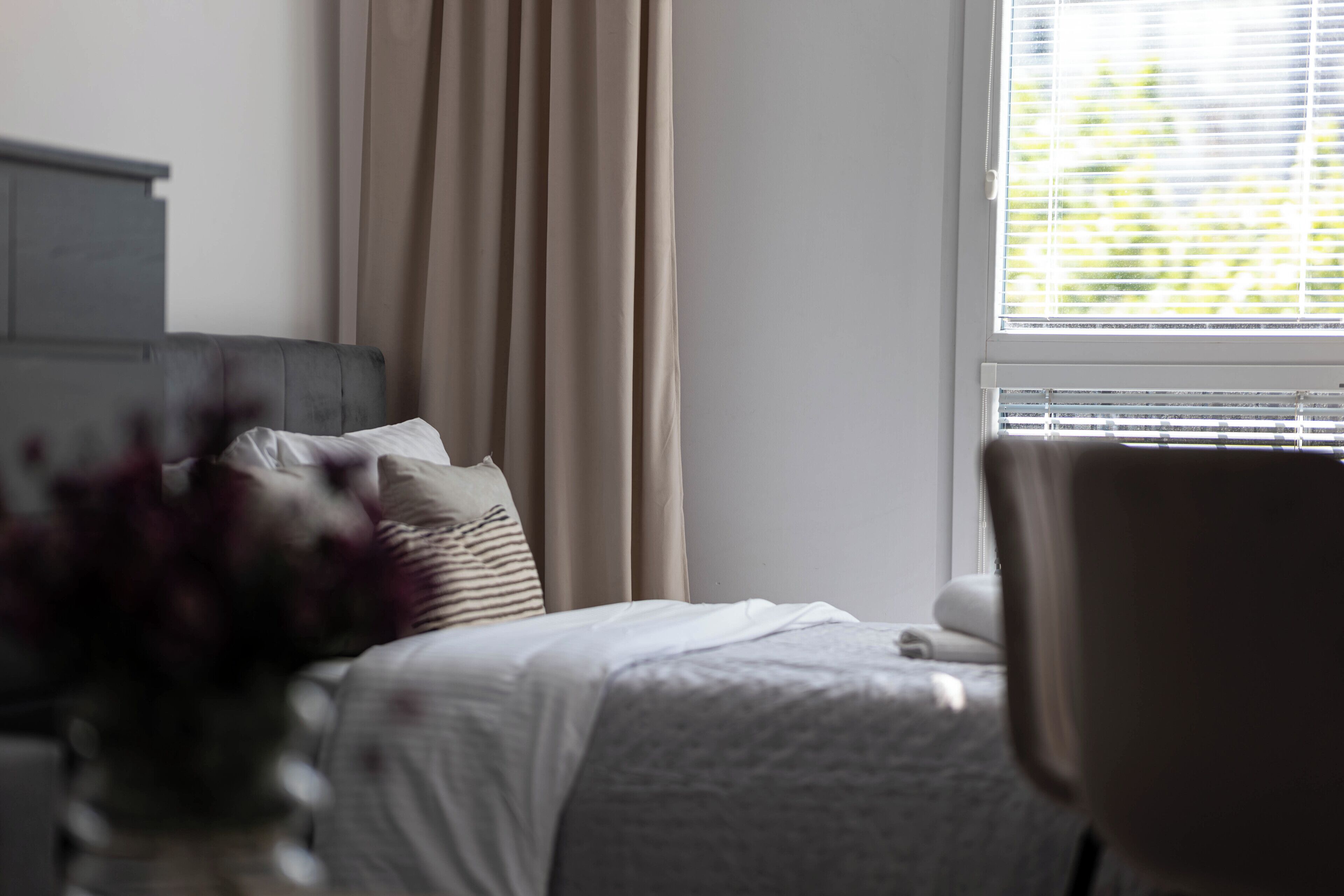 A bedroom with a bed, a chair, a window with blinds, and a curtain.